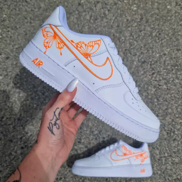 Nike Air Force 1 custom avec deux papillons orange fluo accrochés au swoosh externe entouré d’un halo lumineux orange